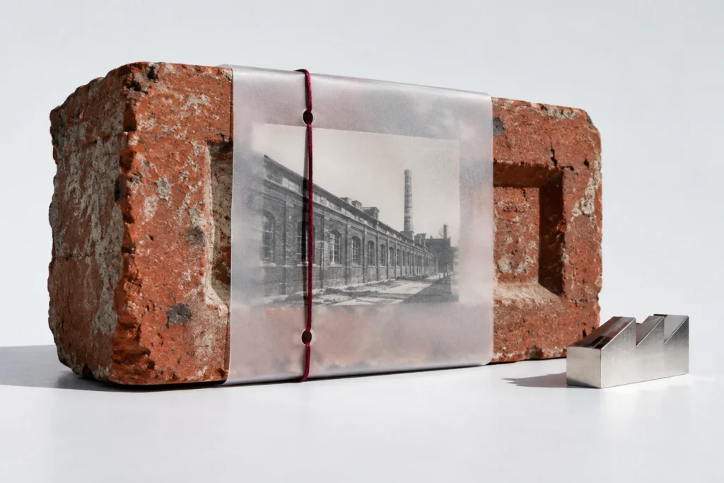 An original red industrial brick wrapped in frosted translucent plastic, holding an imagined warehouse photograph with oxblood thread; a small chrome architectural model sits beside it.